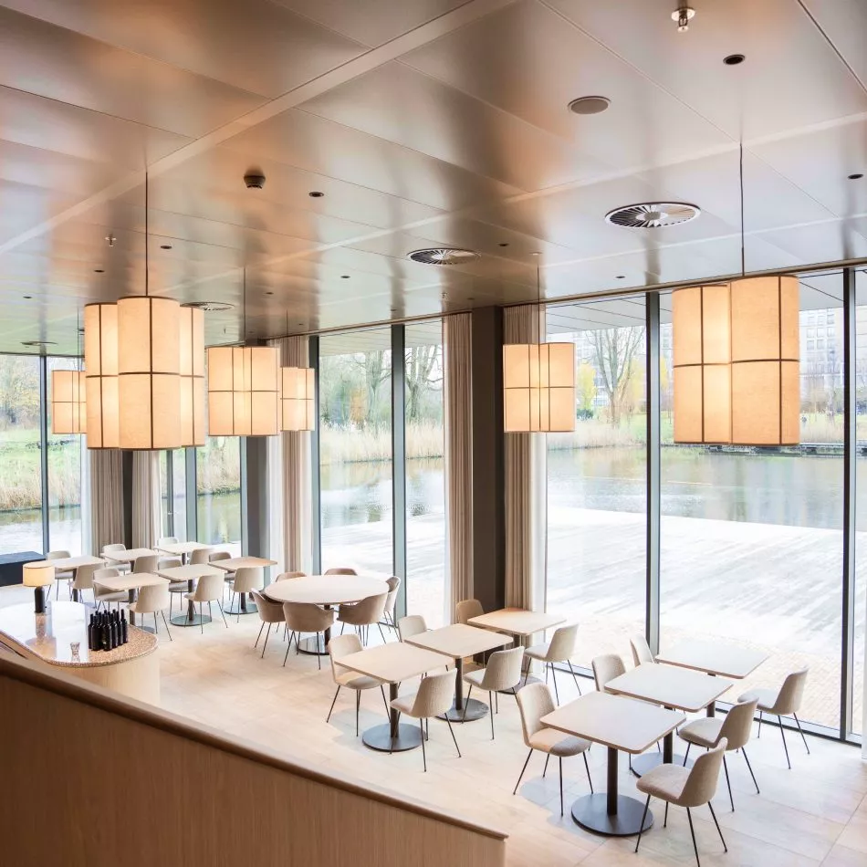 Lichte horecaruimte met grote ramen, moderne hanglampen, beige stoelen en houten tafels met uitzicht op het water.