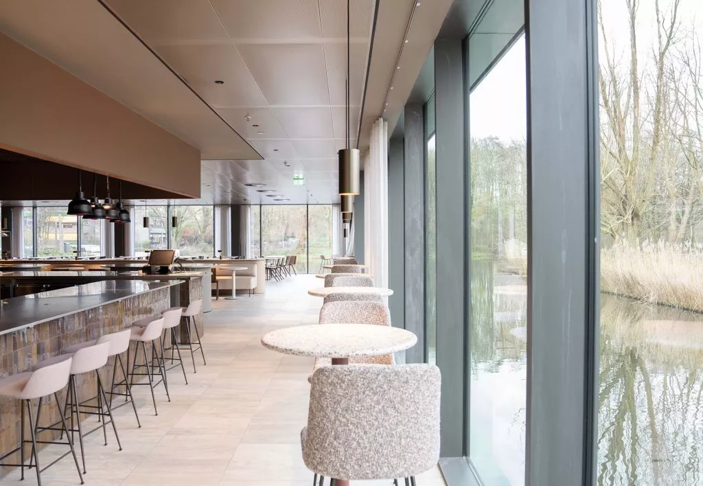 Kantoorruimte van CM.com Amsterdam met hoge ramen langs het water, barkrukken aan hoge tafels en een lichte, moderne interieurafwerking.